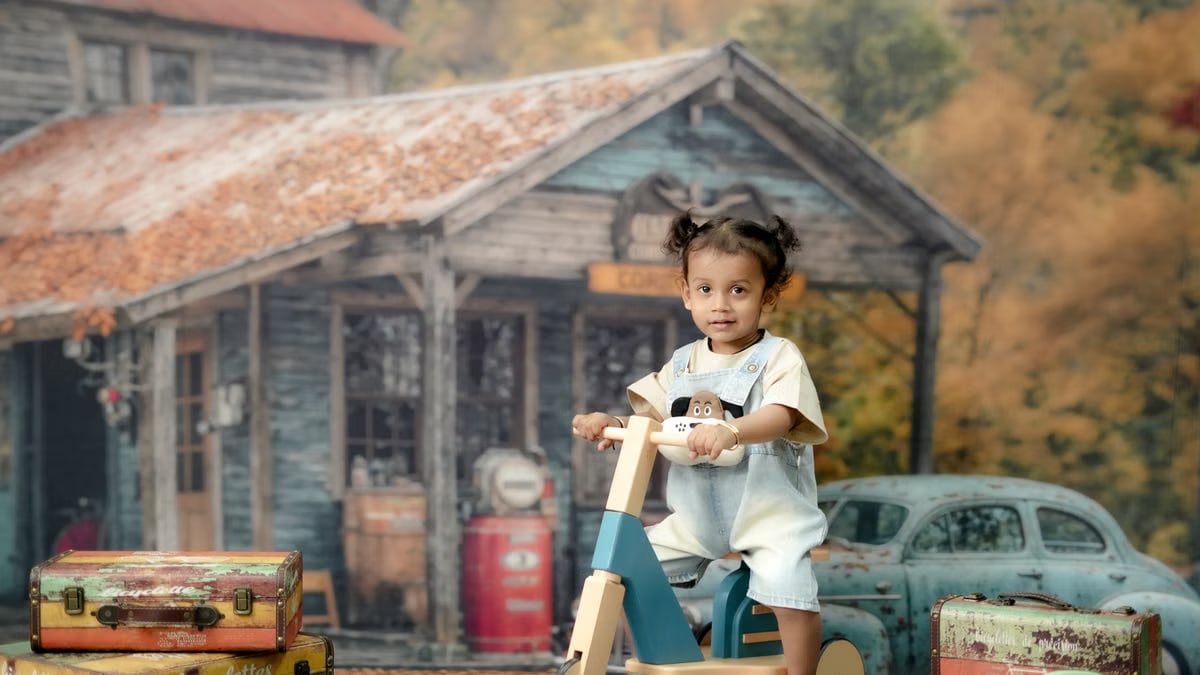 A young child riding a wooden tricycle in front of a rustic autumn-themed backdrop with vintage elements.